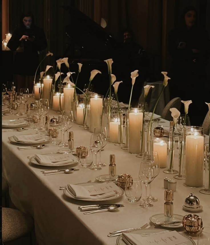 A long dining table set with plates, glasses and tall candles, with a grand piano in the shadows behind